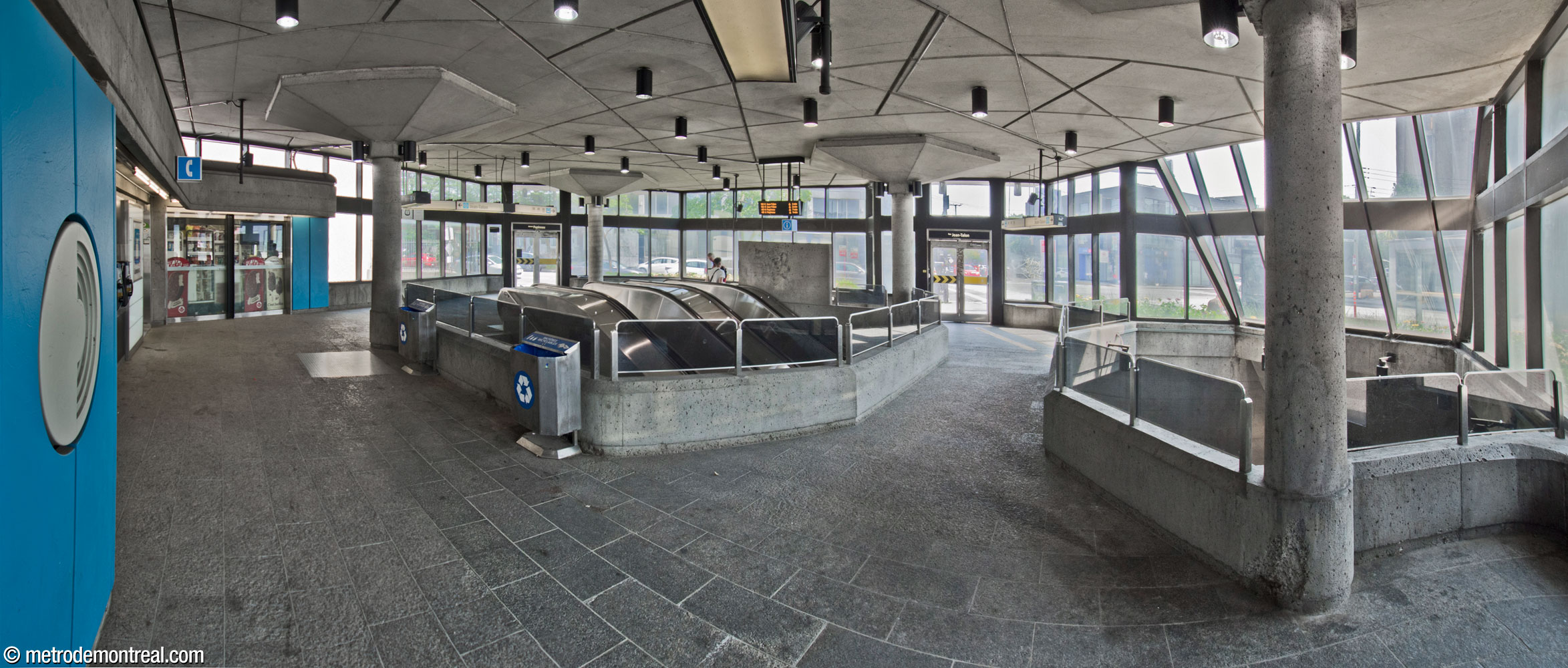 Interior of the eastern headhouse, glazed along most of its perimeter to give it an airy feel. The escalators emerge in the centre of the floor, an effect enhanced by a bridge crossing over the escalator well between the two sides of the headhouse. The sky blue wall panels continue one of the accent colours from the eastern end of the platforms.