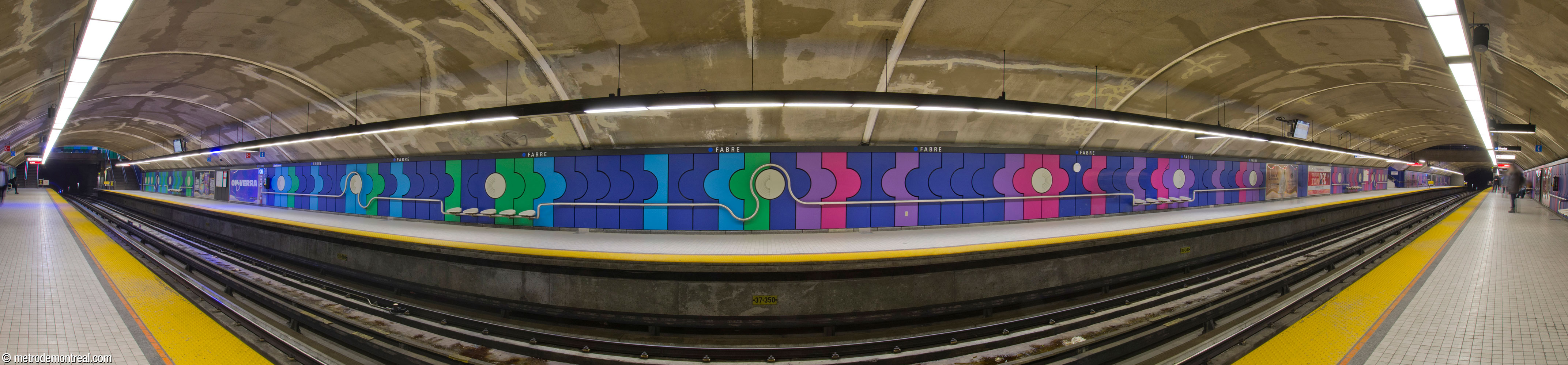 Panorama of the trainroom at Fabre metro station, seen from the Snowdon platform. The trainroom was built in tunnel. The walls are clad in brightly coloured polymer concrete panels, mainly cobalt blue with sky blue and green accents toward the eastern exit (left) and magenta and purple ones toward the western access (right). The panels are punctuated with white discs and a stainless steel rail that curves to highlight the discs and sinks down to incorporate seating. The wall treatments were designed by sculptor Jean-Noël Poliquin.