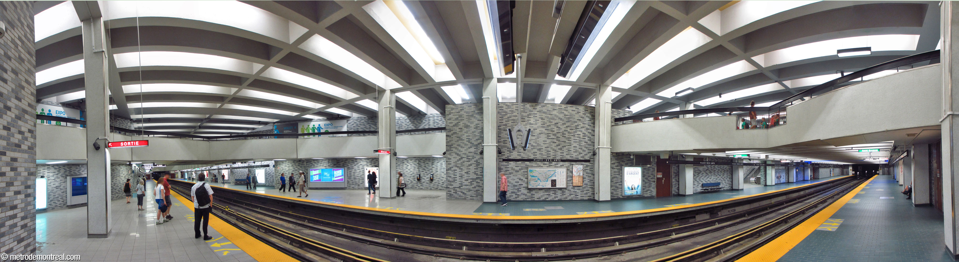 The main volume in the centre of the trainroom, near the staircases leading to the east ticket hall. The broad, impressive volume is defined and overlooked by the parapet of the east fare-paid zone (right) and by a crossover between the overhead walkways between the ticket halls (left). Tall, flat screen walls (centre) separate the stairwells from the volume, and a row of square white pillars along each platform support the roof while further defining the space.