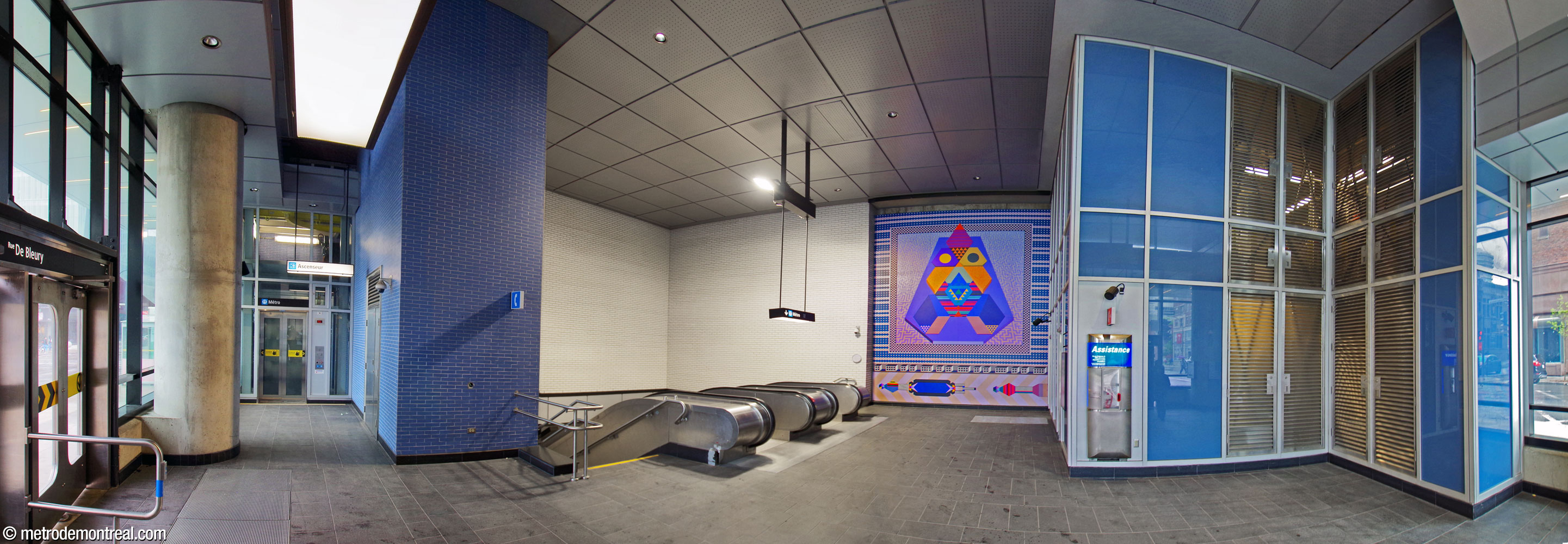 Panorama of the interior of the De Bleury Nord entrance. The stairwell down to the station is at centre-left; the elevator is at left, near the doors. Walls are clad in blue and white bricks. A glass and aluminum wall at right encloses a technical area. Dominique Pétrin’s mural La Réconciliation is on the west wall above the head of the stairs, centre.
