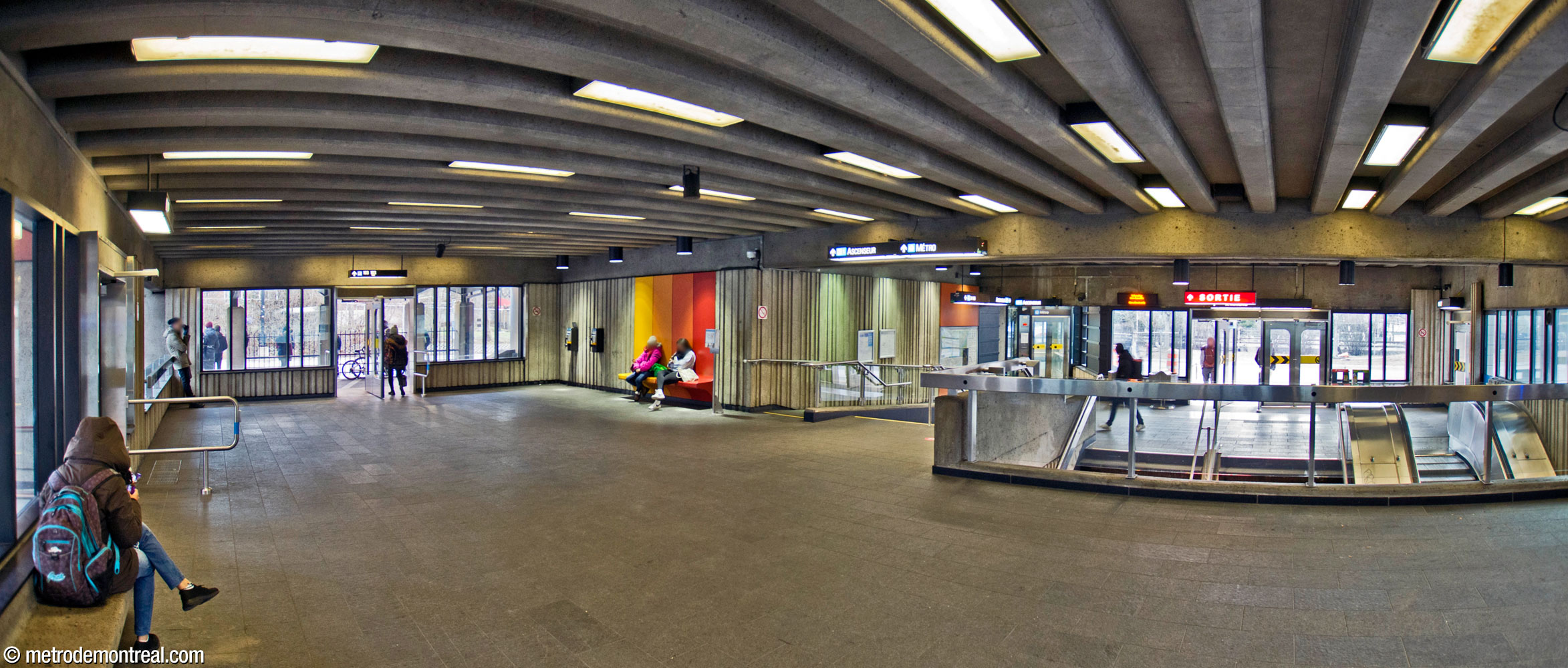 Vue intérieure à partir de la section surélevée à l'arrière de l'édicule. On voit l’ascenseur au fond au centre droit, près de l’entrée principale. Au centre gauche, un banc partage les coloris des sièges au niveau des quais.