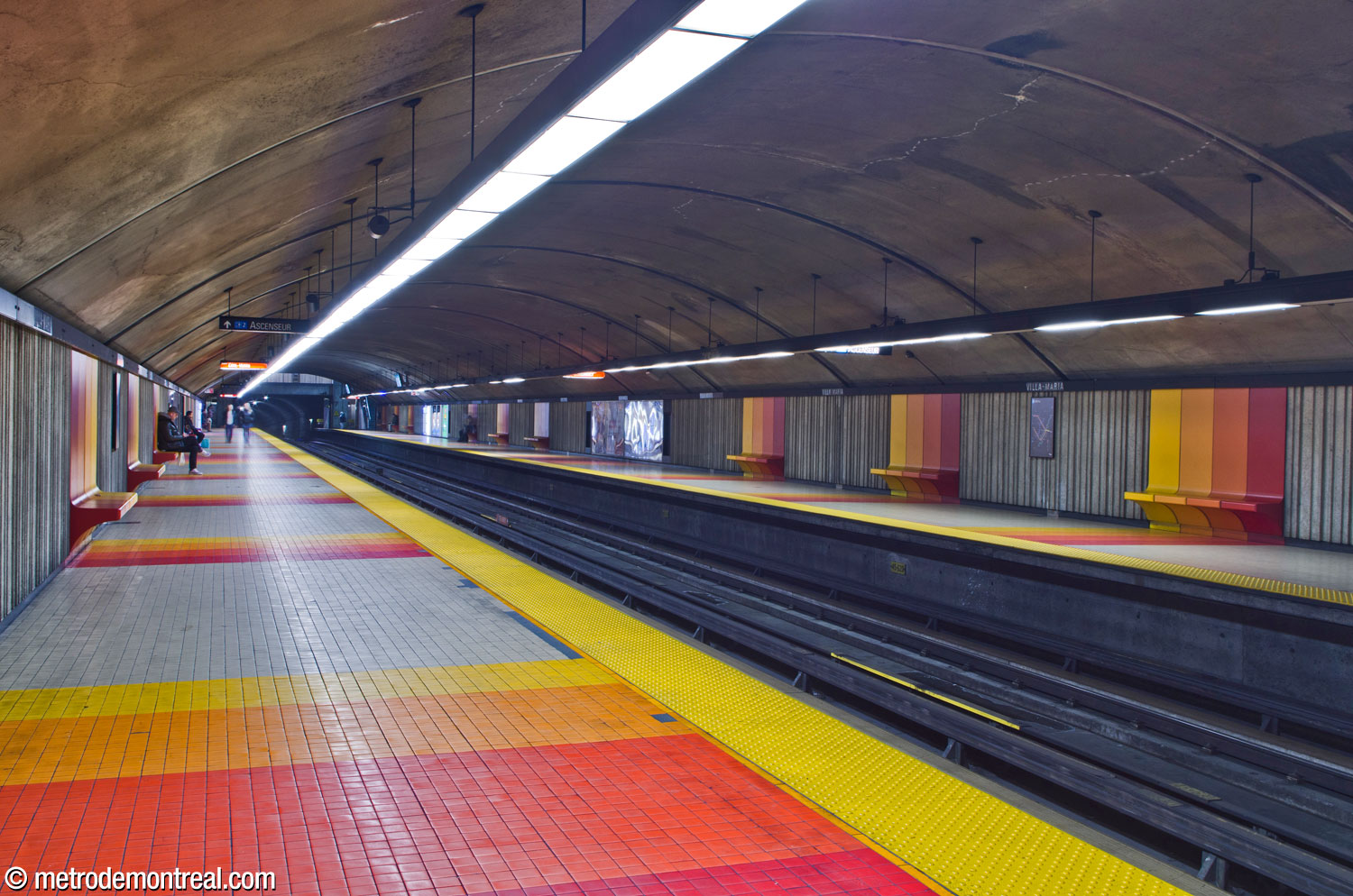 Vue de la nef de la station, creusée dans le roc. On regarde vers le sud à partir du quai Côte-Vertu. Chaque banc présente un dégradé des couleurs jaune, orange, rouge orangé et rouge et est agencé avec des bandes sur le plancher. La couleur jaune se situe du côté plus près du soleil, c'est-à-dire de la sortie.
